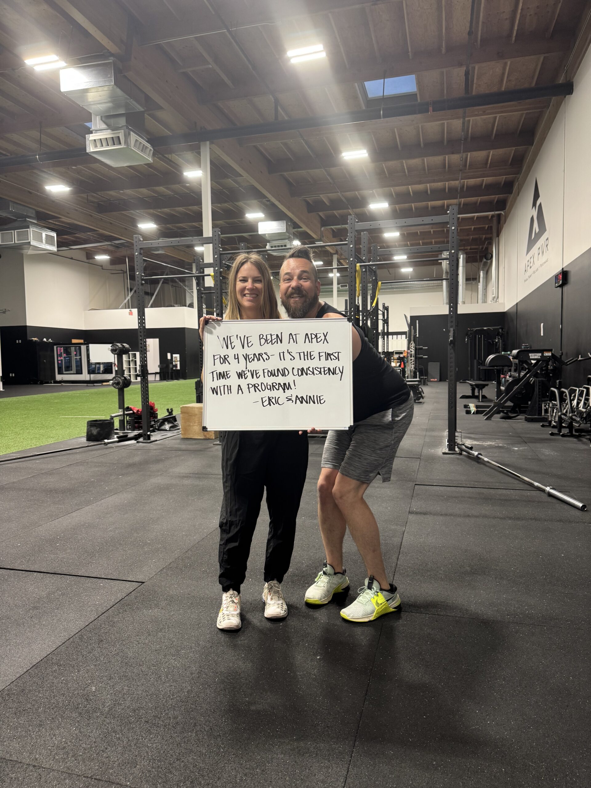 Eric and Annie Wise holding a whiteboard at APEX PWR in Tigard, Oregon, marking 4 years of consistent strength training together with their handwritten note about finding consistency with a program