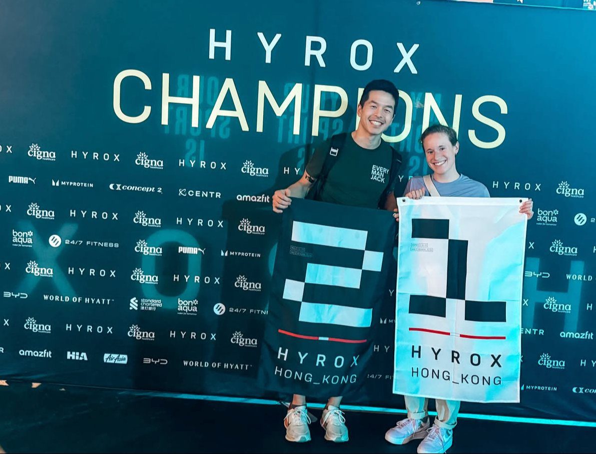 Dr. Kevin Le, DPT and Batya Beard at the Hyrox Champions backdrop, Hyrox Hong Kong 2026, holding their 3rd place and 1st place podium flags
