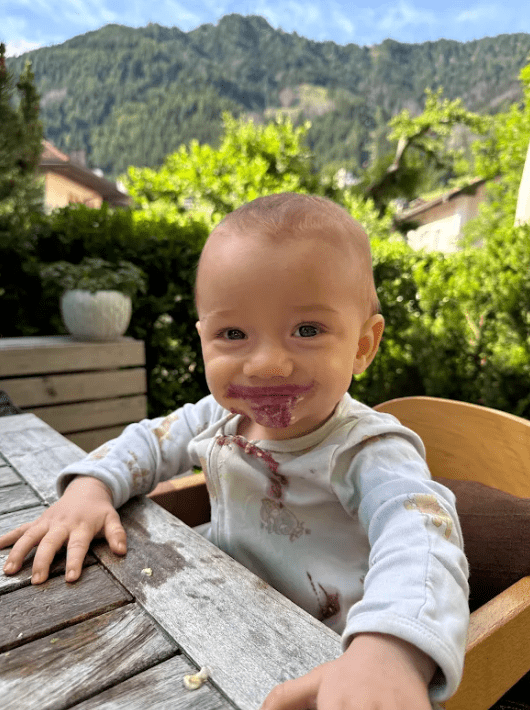 Oslo, Jennie Carolan's son, smiling in a high chair at an outdoor restaurant in the Swiss Alps during the family's European trip