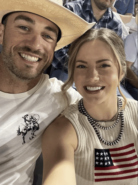 Colin Feldtman, CSCS, in an APEX PWR shirt and St. Paul Rodeo cap, holding his young daughter with his wife at the rodeo grounds over the 4th of July